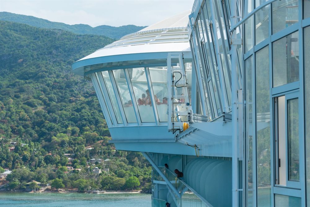Glass-enclosed observation area on a cruise ship above a green coastline